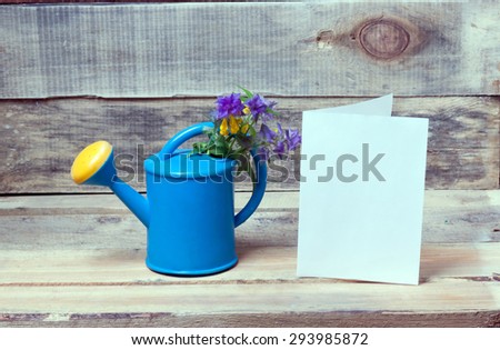 watering can, flowers and a blank sheet of paper on a wooden bench.clouseup