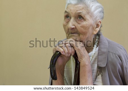 sad old lady's portrait with a walking stick  in front of a light brown wall