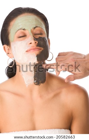 Hand of cosmetology professional beautician applying facial masks of different color on face of ethnic woman wrapped in bath towel during beauty treatment session, relaxed with closed eyes