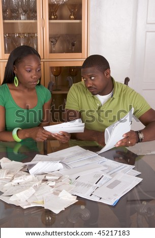 Young black African American couple sitting by glass table and working through pile of bills, frustrated by amount of expenses during economic crises recession times hoping for stimulus plan to work