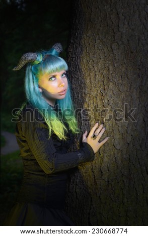 Beautiful woman with color hair and horns in the forest holds a tree