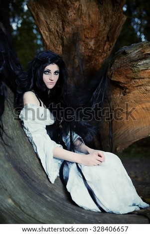 Young beautiful witch with long dark hair in root of old tree