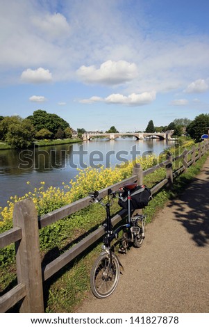 Folding bicycle stands alone near River Thames, Hampton Court Bridge in the distance.