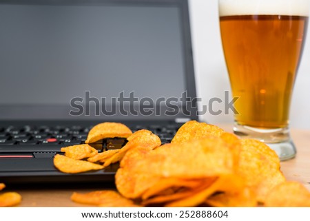 closeup of open laptop with chips scattered on keyboard and glass of beer in background - stock photo