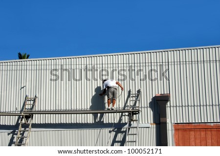 Man works from high scaffolding spray painting the exterior of a warehouse on the Big Island of Hawaii.  He is wearing no safety belt as he works.