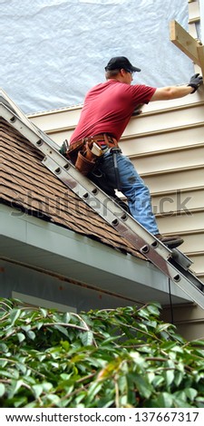Young man perches on metal ladder on roof top.  He is installing siding on his home.  He is wearing a tool belt, red shirt and work jeans.