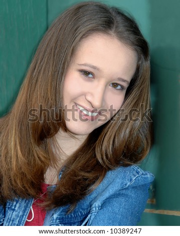 Female teen leans against a green wooden wall.  She has a gentle, sweet smile on her face.  She is wearing denim.