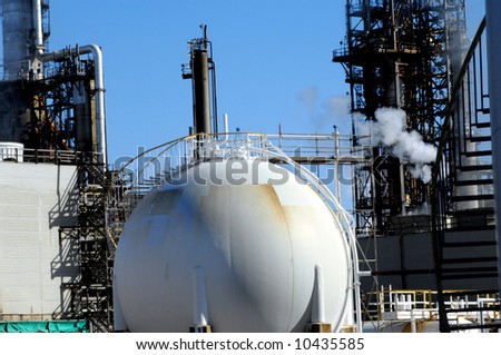 Storage tank is round and white with ladder running up side.  Industrial scene in background complete with steam and stack.