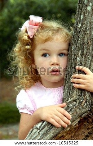 Young girl peeks out from behind a tree.  She has golden curls and a pink bow.