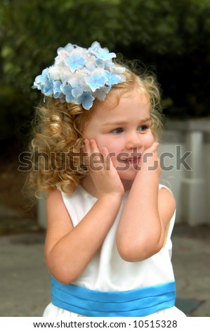 Young girl plays peek-a-boo from behind her hands.  She has long golden curls and has flowers in her hair.