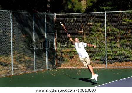 Young male tennis player gives extreme effort and returns a high tennis ball.  Feet are off ground and his stretch is to his limit.