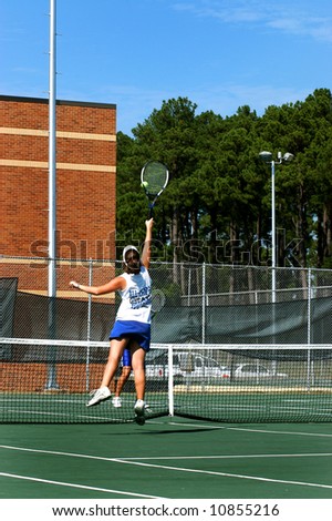 Female tennis player jumps high to retrieve a tennis ball while playing the net at a college campus.  Blue skies.