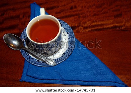 Background has triangle folded bright blue napkin.  Cup of amber hot tea is hot and ready in a blue patterned china cup and saucer.