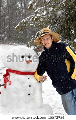 Clowning around with the snowman, young man borrows hat and pipe and leans against snowman's shoulder.