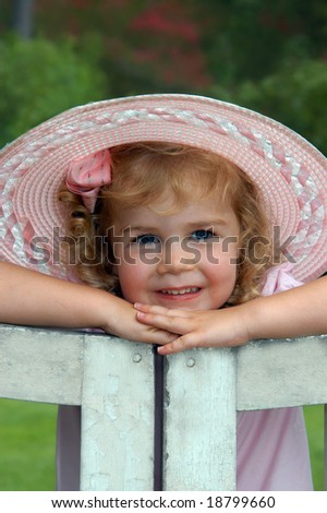 Beautiful little girl poses on a garden gate.  She is wearing a pink, hat, bow and dress.
