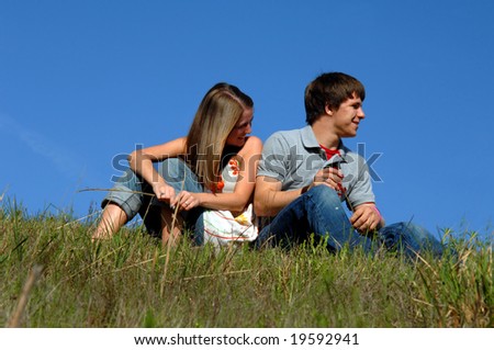 Blue skies frame young teen couple.  They are sitting on a grassy hill side-by side.