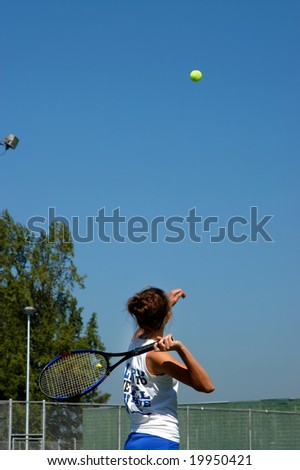 Female teen throws ball into air and swings racket back for serve.  Blue sky.