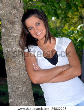 Female teen leans her shoulder against a tree and crosses her arms.  She is smiling and wearing a white shirt.