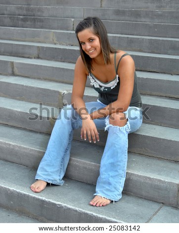 Happy teenager sits on concrete steps of high school.  She is wearing worn jeans and spaghetti strap top.