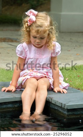 In a world of her own, small blond haired child wet her feet in pool.  Pink dress and hairbow.