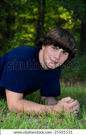 Enjoying a day outdoors, male teen lays on grass with woods behind him.  He is wearing a navy blue tee shirt.