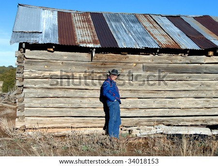 Man wearing jeans and a cowboy hat, leans against the wood and adobe wall of an abandoned cabin.  Building is outside of Alberquerque, New Mexico.
