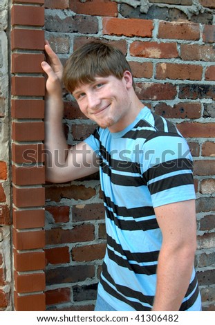 Young man leans against an old brick wall and the beginnings of a smile are forming.  He has on a blue and black striped shirt.