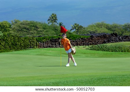 Woman stands besides hole marked by a red flag on a golf course on the Big Island of Hawaii.  Mauna Loa rises in the background behind golf course.
