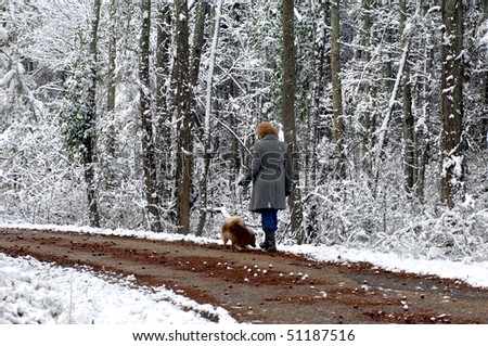 Woman and her pomeranian ambles down a quiet backroad surrounded by snow covered trees.