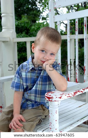 Small boy sits in a white wooden rocking chair on his front porch.  He is leaning on his elbow and waits patiently for his family.