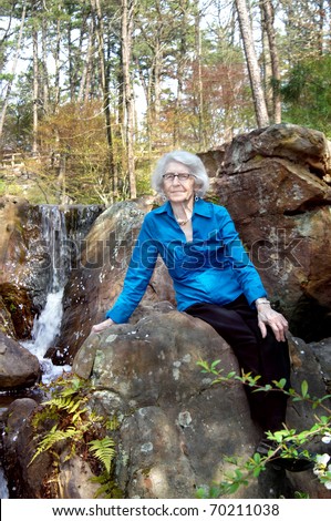 Elderly woman sits besides waterfall at Garvin Woodland Garden in Hot Springs, AR.  She is wearing a vivid blue shirt.