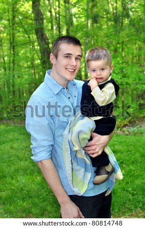 Young man holds little boy surrounded by green forest.  Man is smiling and toddler is chewing on his finger.
