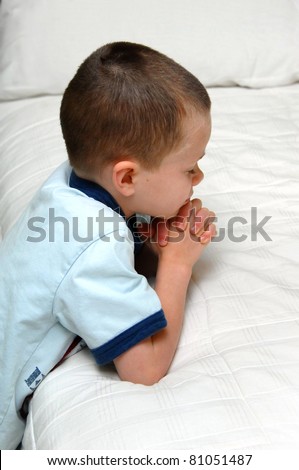 Small child kneels besides his bed and folds his hands in prayer.  He is wearing a blue shirt and kneeling besides a white covered bed.