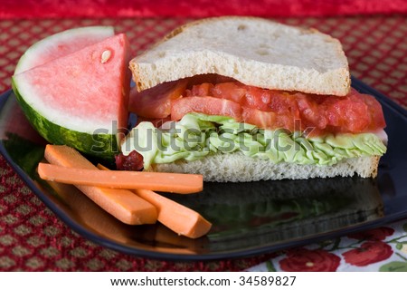 Tomato and butter lettuce sandwich on a black plate with carrot sticks and watermelon slices.