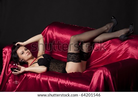 Sensual woman in sexy black lingerie sitting on a bed covered in satin, enjoying a glass of wine. Studio shot