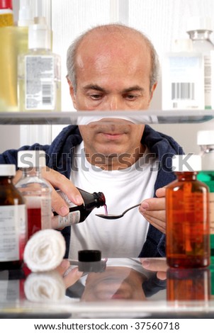 A sick man pouring cough syrup into a spoon viewed through a medicine cabinet