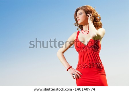 high end fashion shot of sexy red hair girl wearing red dress with vibrant clear blue sky in background in sunny day
