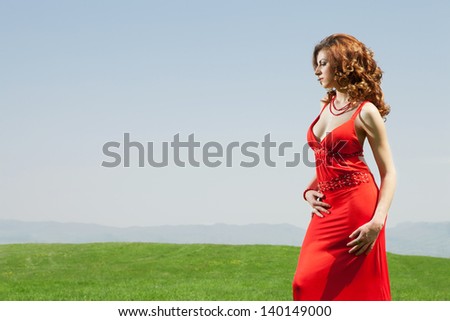 high end fashion shot of sexy red hair girl wearing red dress in complementary green grass field in sunny day
