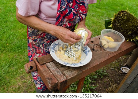 senior woman with apron hands grate peel potatoes with steel shredder tool on wooden outdoor table.