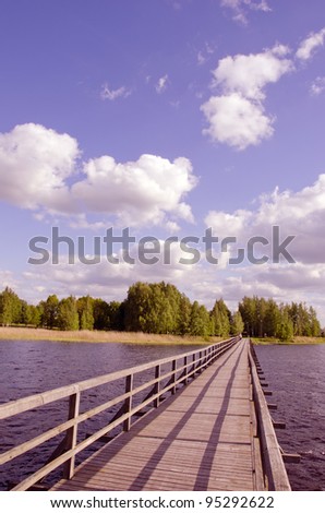 Long wooden footbridge with handrails over the lake and the man walking far away. Forest in the distance and the cloudy sky.