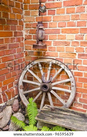 Antique wooden carriage wheel with binding of metal and horseshoe on it. Kerosine paraffin rusty lamp. Red brick wall. Fern leaves.