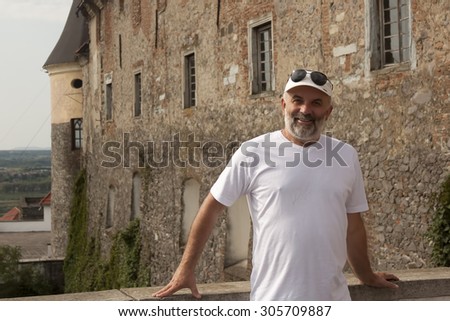 Portrait of a middle-aged man on a background of a wall of an old castle in the white T-shirt and a baseball cap and sunglasses on his forehead