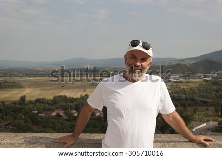 Portrait of a middle-aged man in the background fields and low mountains in white T-shirt and a baseball cap and sunglasses on his forehead