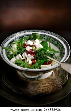 Creatively lit fresh and healthy Greek feta cheese and pomegranate rocket salad against a dark rustic background with copy space. Concept image for a restaurant menu cover design.