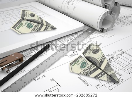Paper airplanes of dollars lying on laptop keyboard and engineering drawings. Tools are close by. Concept of industry and business