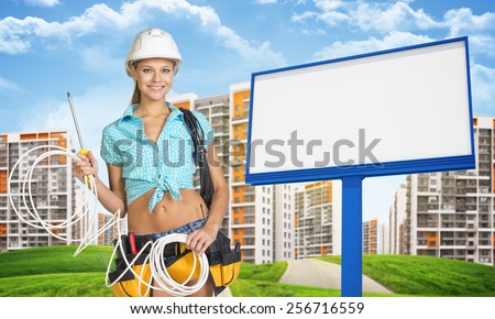 Woman in hard hat and tool belt, holding coils of cable, standing among green hills by the road and blank billboard, looking, at camera, smiling. High-storey buildings as backdrop