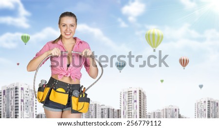 Woman in tool belt with different tools connects two flexible hoses, smiling. Buildings with air balloons as backdrop