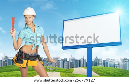 Woman in helmet and tool belt with different tools holding wrench and two flexible hoses, smiling. Green hills with road, buildings and empty billboard as backdrop