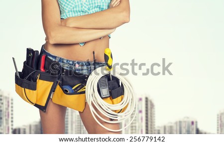 Woman in tool belt with different tools standing crossed arms. Cropped image. Buildings as backdrop