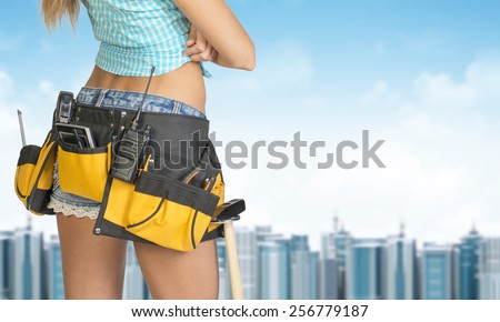 Woman in tool belt with different tools stands back. Cropped image. Building and sky as backdrop
